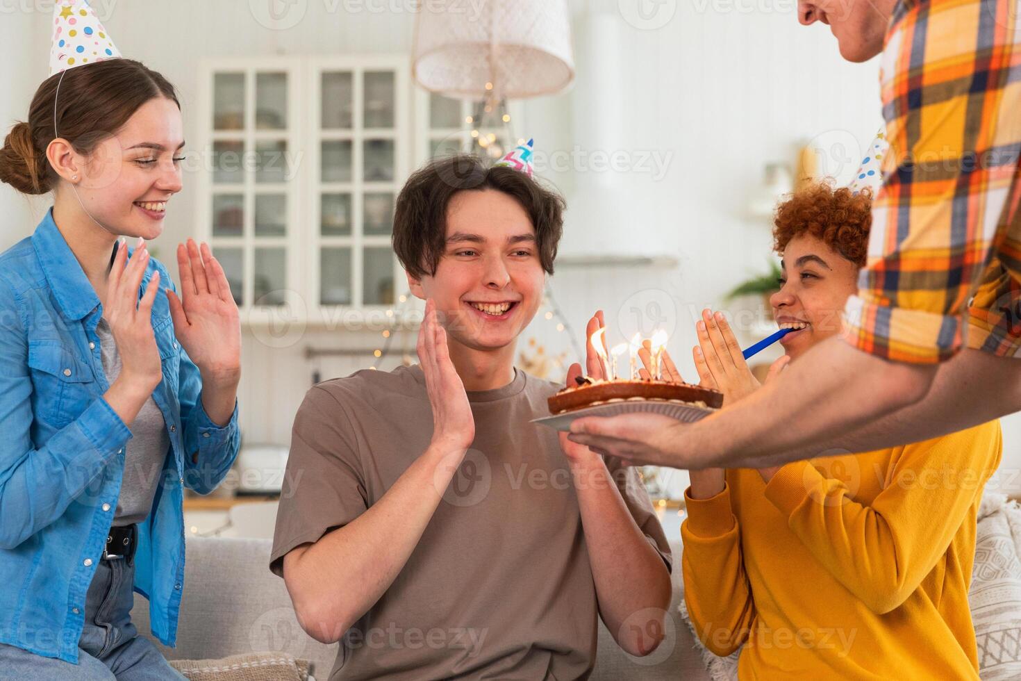 Make a wish. Man wearing party cap blowing out burning candles on birthday cake. Happy Birthday party. Group of friends wishes guy happy birthday. People celebrating birthday with party at home photo