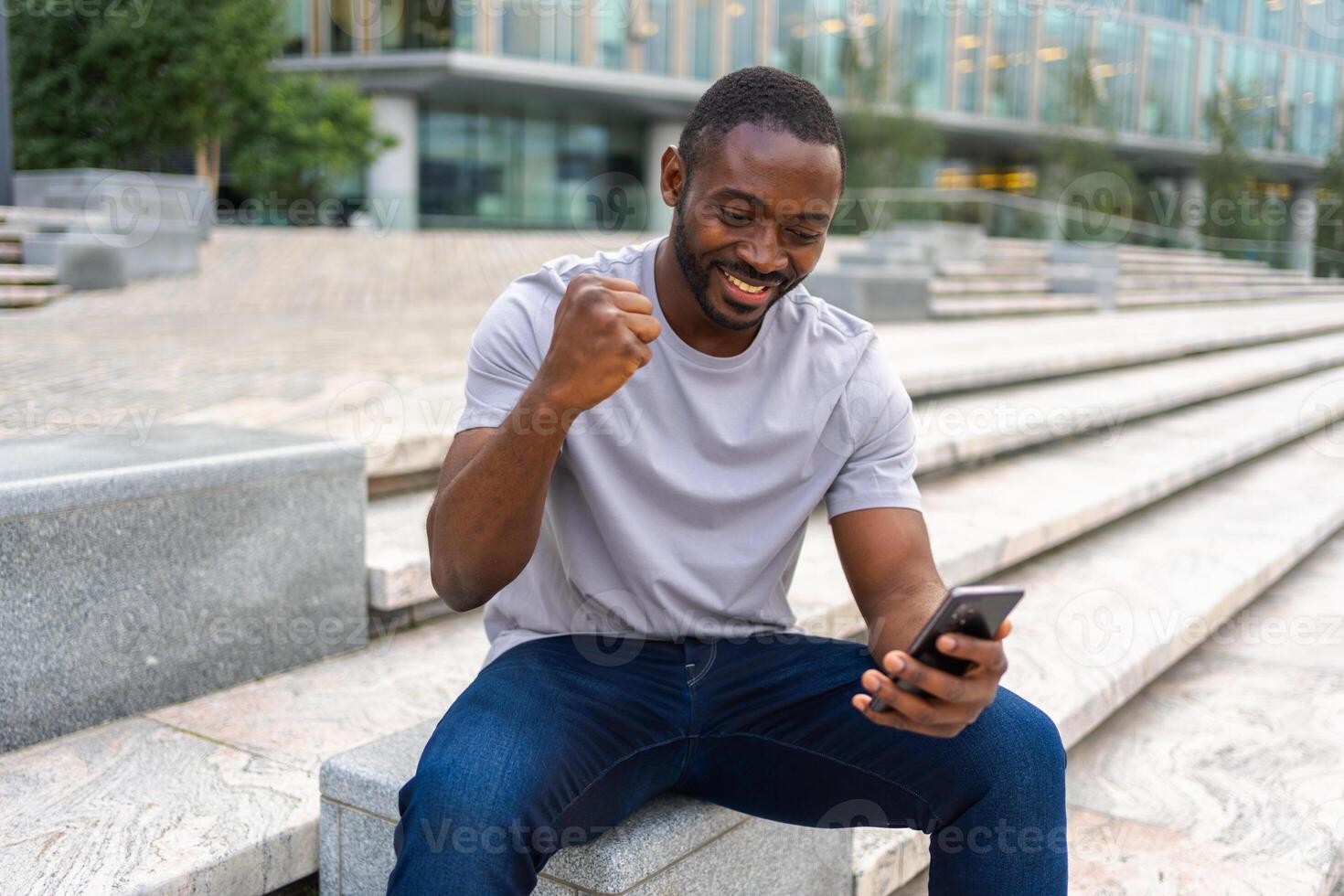 Happy African American man euphoric winner with smartphone on street in city. Person guy looking at cell phone reading great news getting good result winning online bid feeling amazed. Winning gesture photo