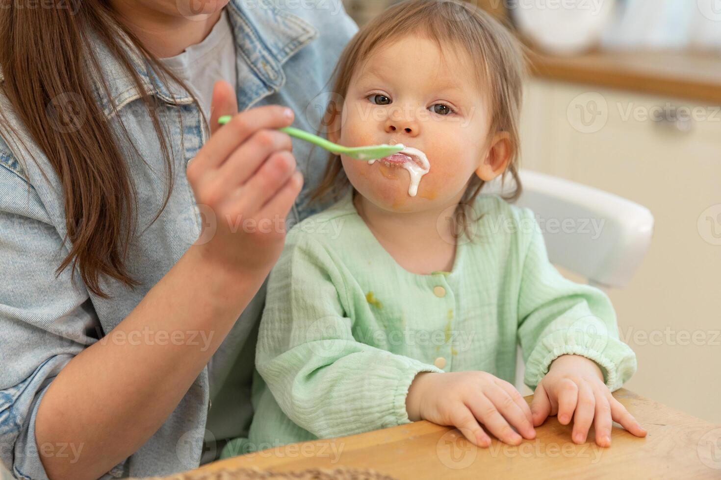 contento familia a hogar. madre alimentación su bebé niña desde cuchara en cocina. pequeño niñito niño con sucio gracioso cara come sano comida a hogar. joven mujer mamá dando comida a niño hija foto