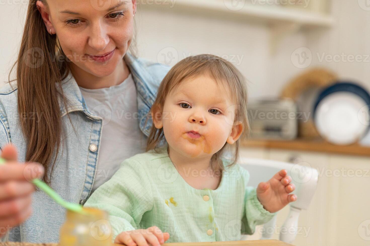 Happy family at home. Mother feeding her baby girl from spoon in kitchen. Little toddler child with messy funny face eats healthy food at home. Young woman mom giving food to kid daughter photo