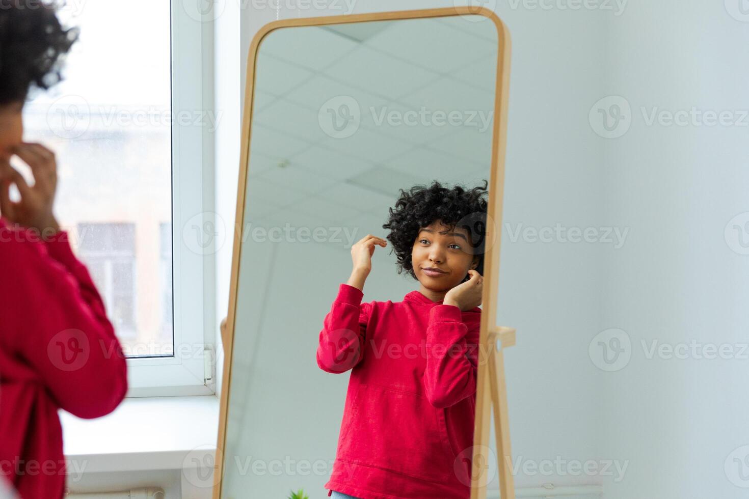 Love yourself. Beautiful young smiling african american woman dancing touching curly hair enjoying her mirror reflection. Black lady looking at mirror looking confident and happy. Self love concept photo