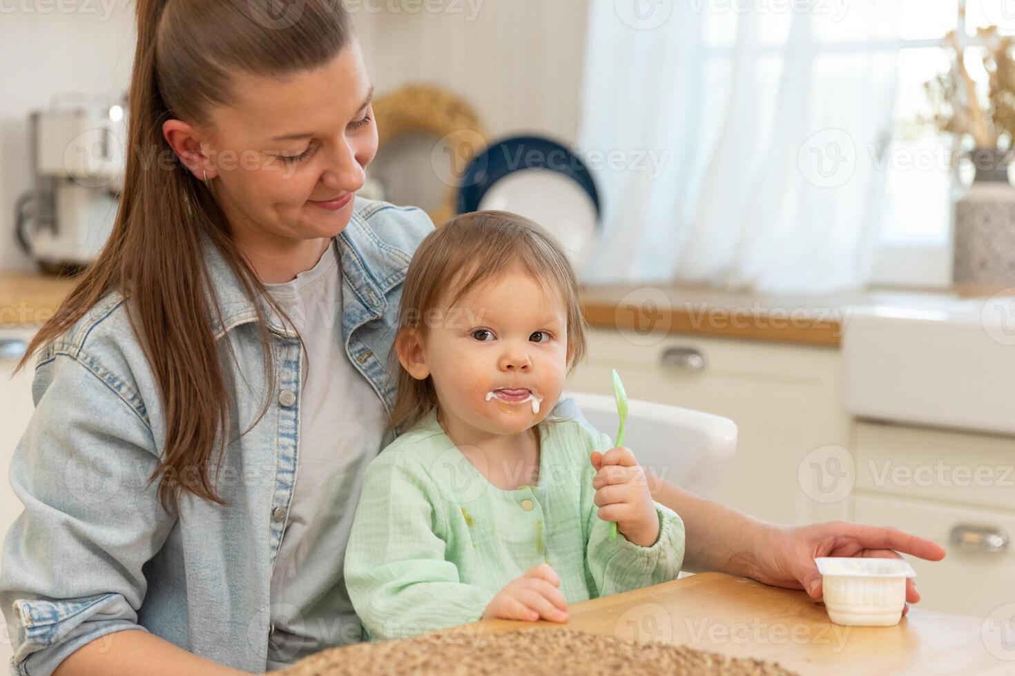 contento familia a hogar. madre alimentación su bebé niña desde cuchara en cocina. pequeño niñito niño con sucio gracioso cara come sano comida a hogar. joven mujer mamá dando comida a niño hija foto