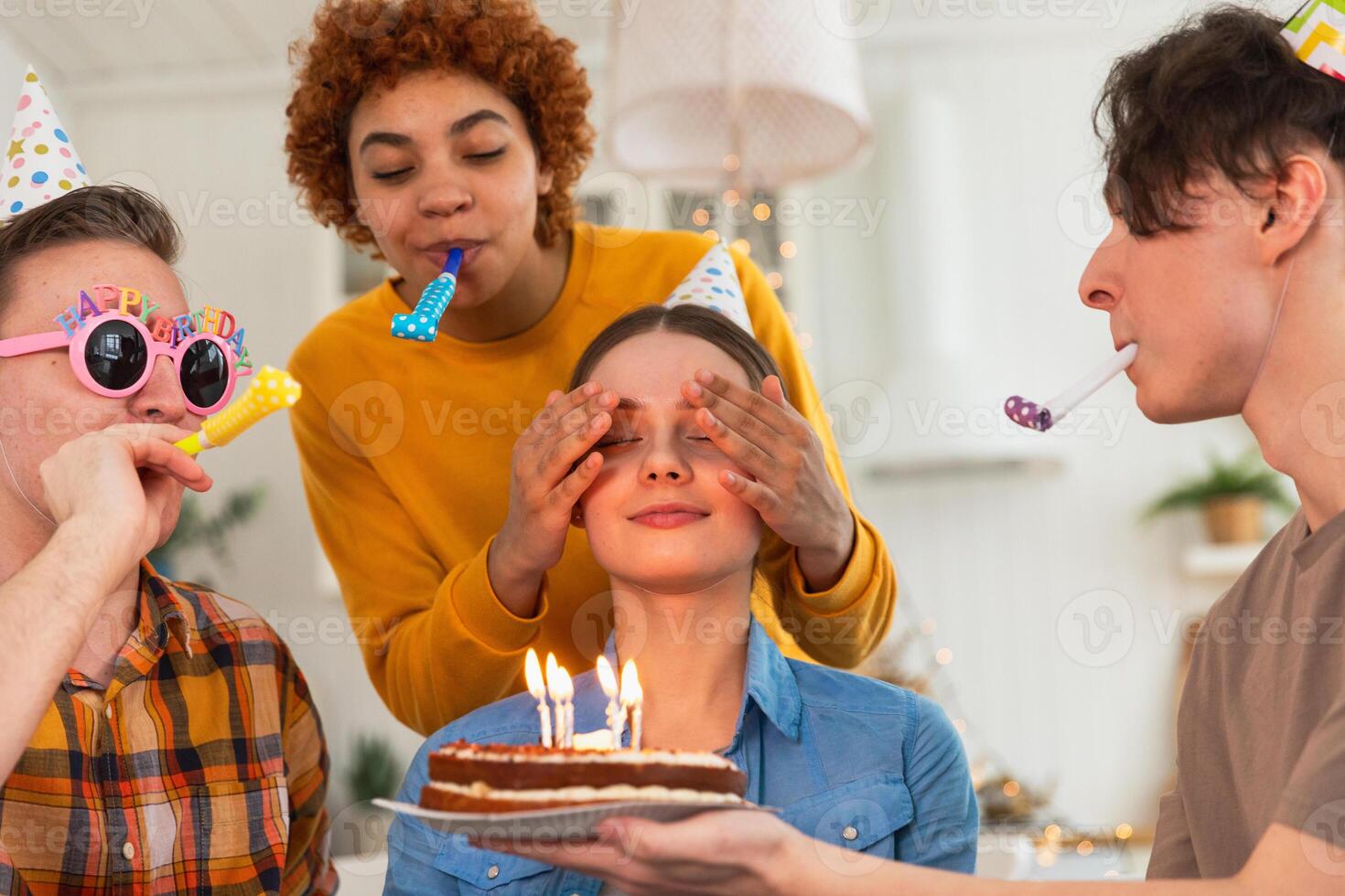 Make a wish. Woman wearing party cap blowing out burning candles on birthday cake. Happy Birthday party. Group of friends wishes girl happy birthday. People celebrating birthday with party at home photo