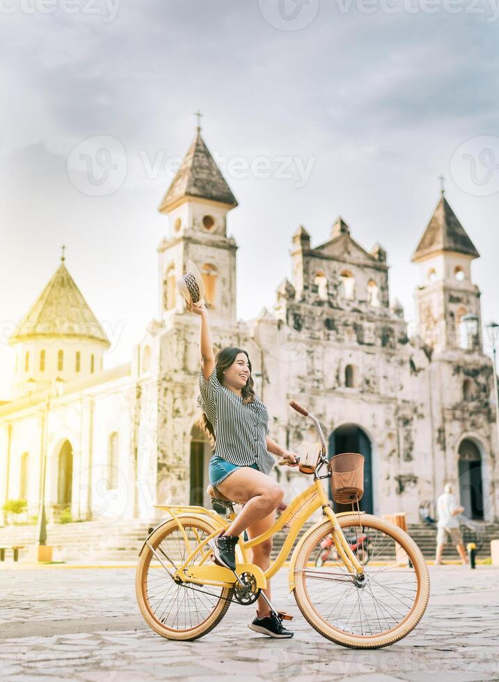 Happy young woman in hat riding a bicycle on the street. Granada, Nicaragua. Lifestyle of a happy girl riding a bicycle holding her hat on the street. Tourism concept photo