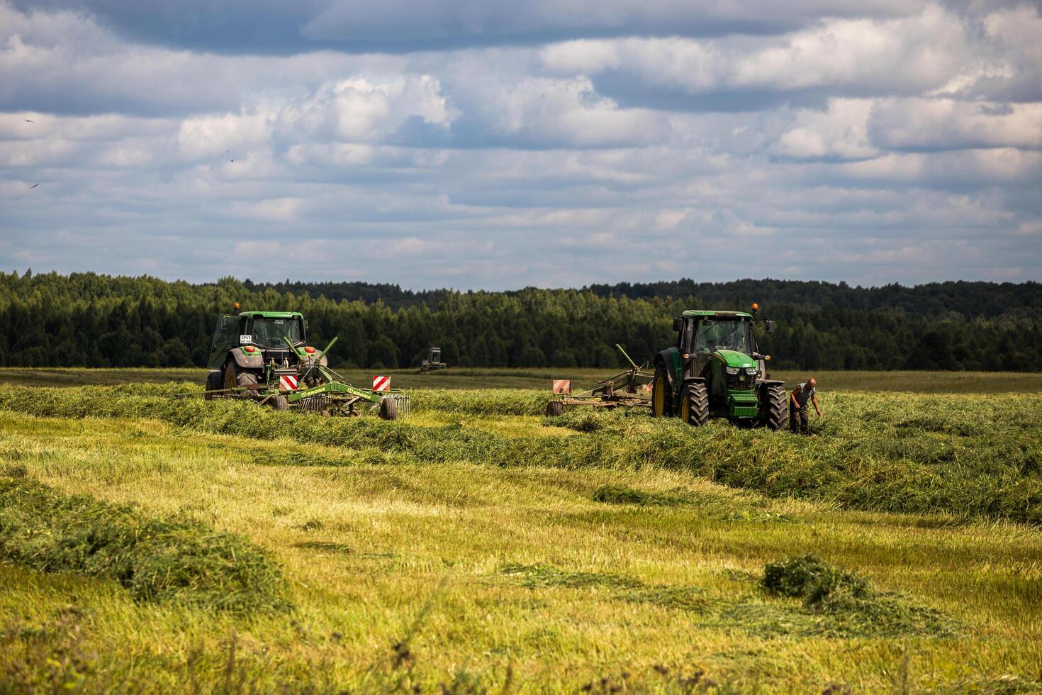 two green haymaking John Deere tractors with Krone plow on summer field before storm - telephoto shot with selective focus photo