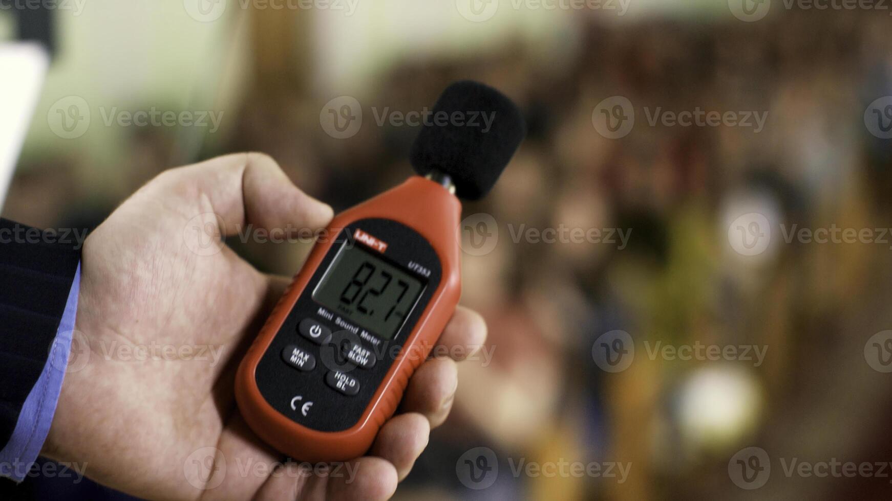 Close up of a man hand holding a red sound level meter and analyzer on clapping audience background. Stock footage. Man measuring the noise level on blurred crowd background. photo