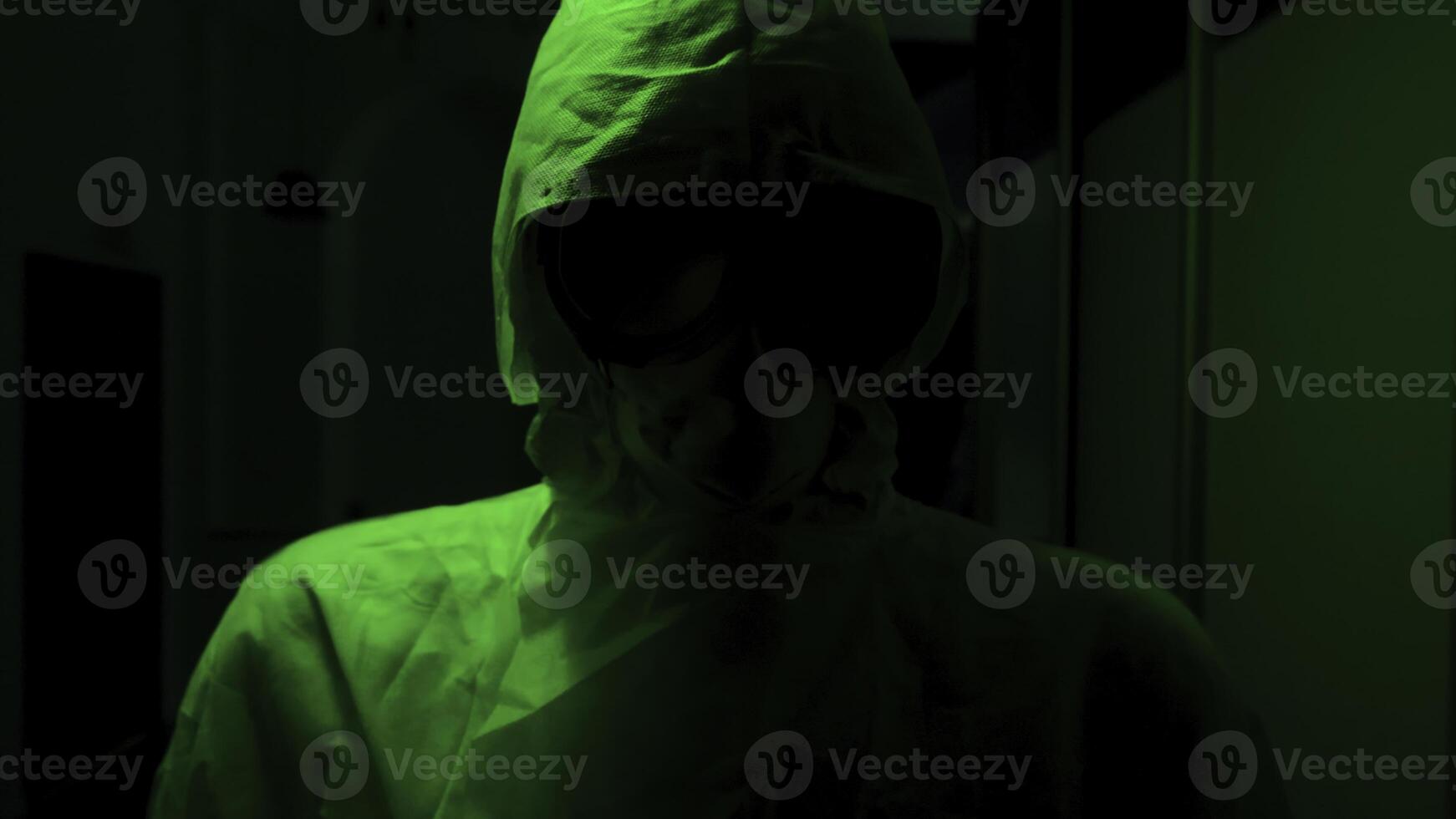 Man in protective suit and mask stands in dark. Stock footage. Creepy doctor in antiviral suit stands in dark corridor with green light. Dangerously high levels of infection photo