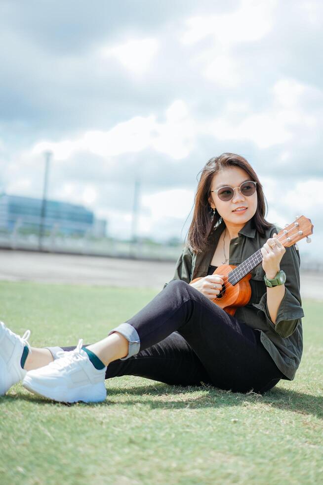 Playing Ukulele of Young Beautiful Asian Woman Wearing Jacket And Black Jeans Posing Outdoors photo