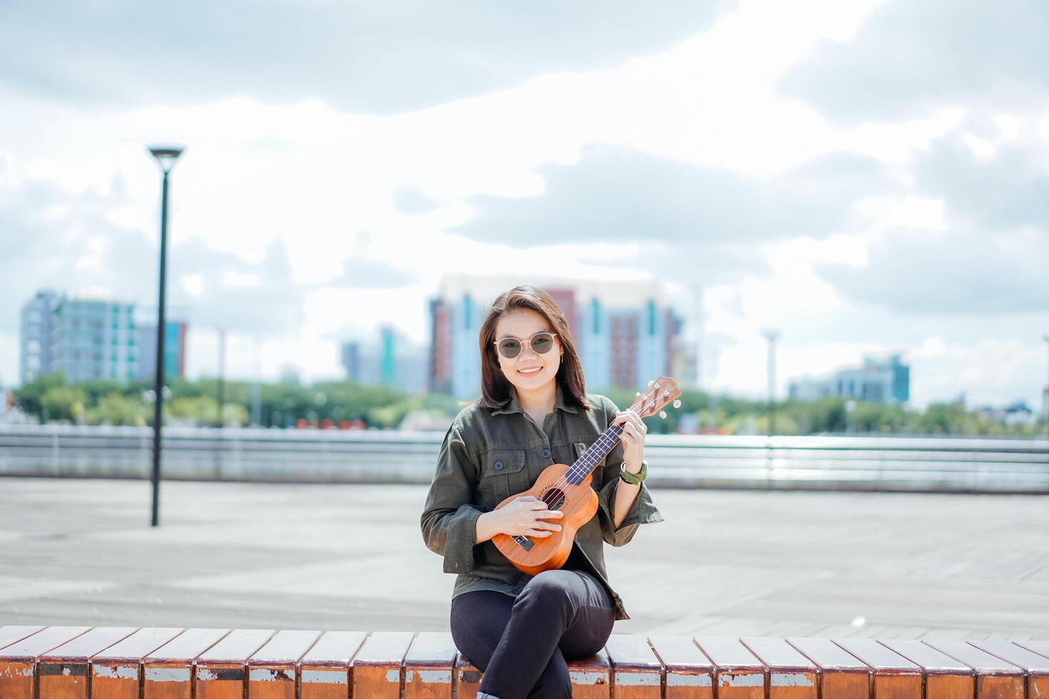 jugando ukelele de joven hermosa asiático mujer vistiendo chaqueta y negro pantalones posando al aire libre foto