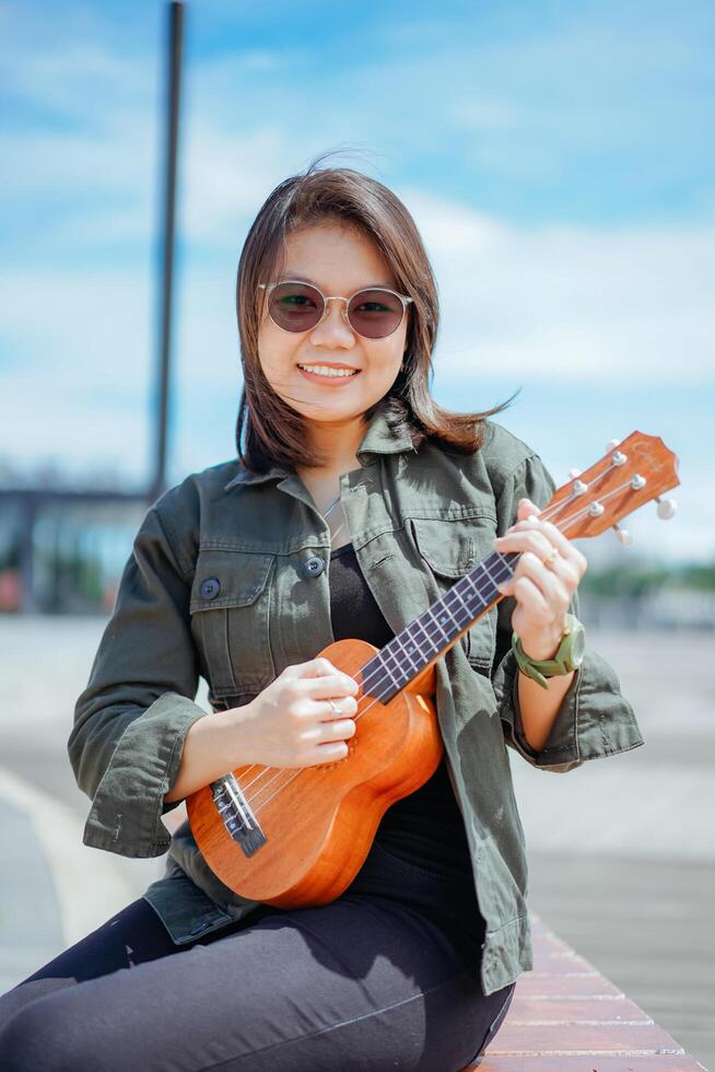 Playing Ukulele of Young Beautiful Asian Woman Wearing Jacket And Black Jeans Posing Outdoors photo