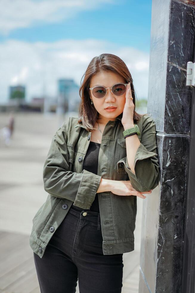 joven hermosa asiático mujer vistiendo chaqueta y negro pantalones posando al aire libre foto