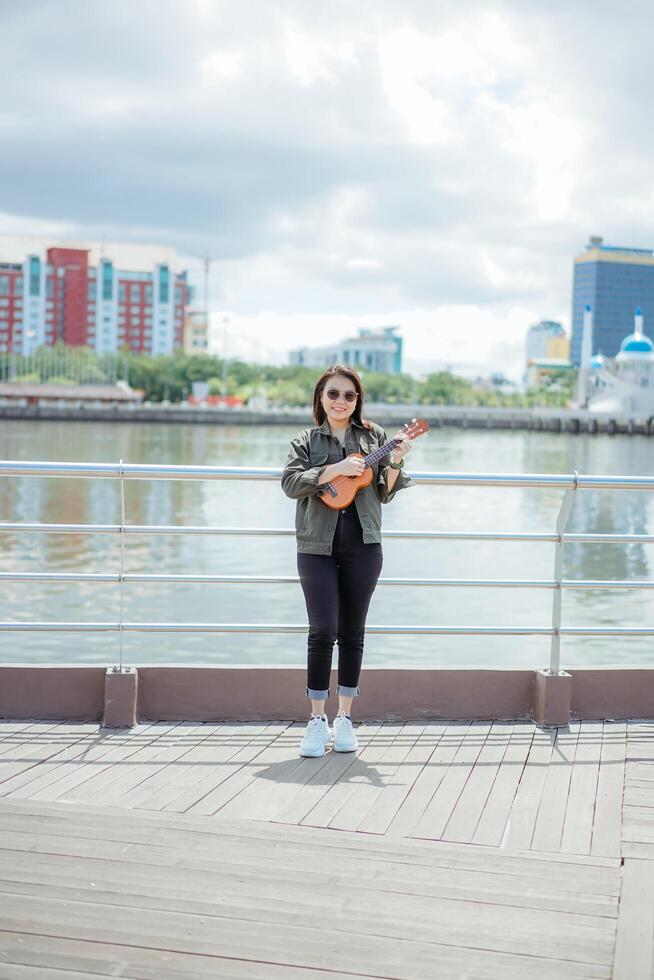 Playing Ukulele of Young Beautiful Asian Woman Wearing Jacket And Black Jeans Posing Outdoors photo