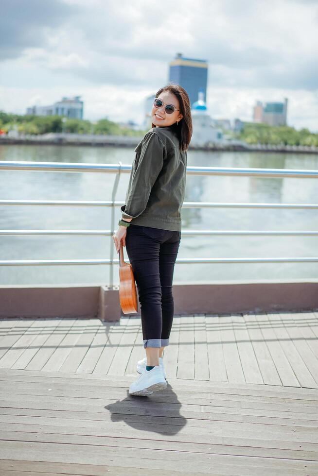 jugando ukelele de joven hermosa asiático mujer vistiendo chaqueta y negro pantalones posando al aire libre foto