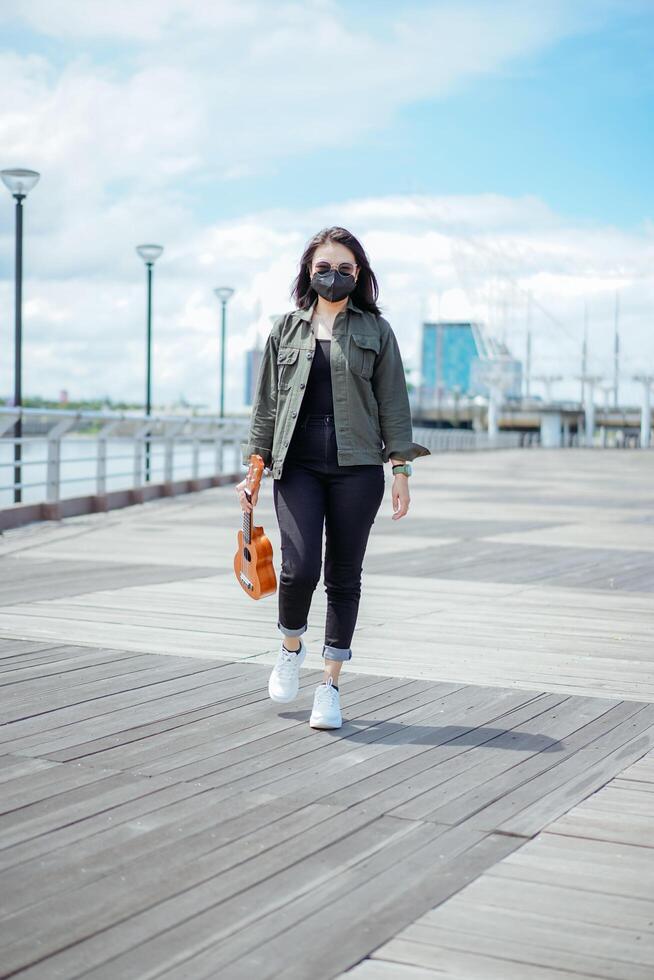 Playing Ukulele of Young Beautiful Asian Woman Wearing Jacket And Black Jeans Posing Outdoors photo