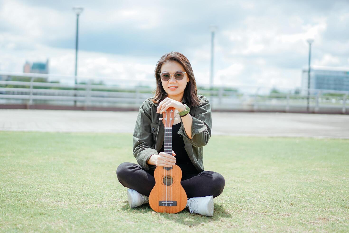 jugando ukelele de joven hermosa asiático mujer vistiendo chaqueta y negro pantalones posando al aire libre foto