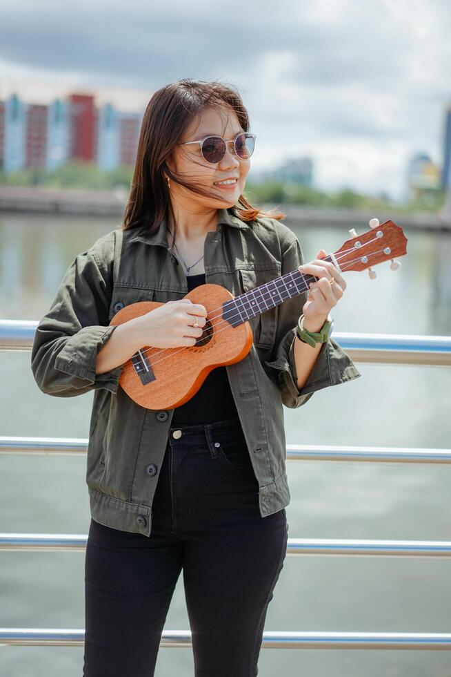 jugando ukelele de joven hermosa asiático mujer vistiendo chaqueta y negro pantalones posando al aire libre foto