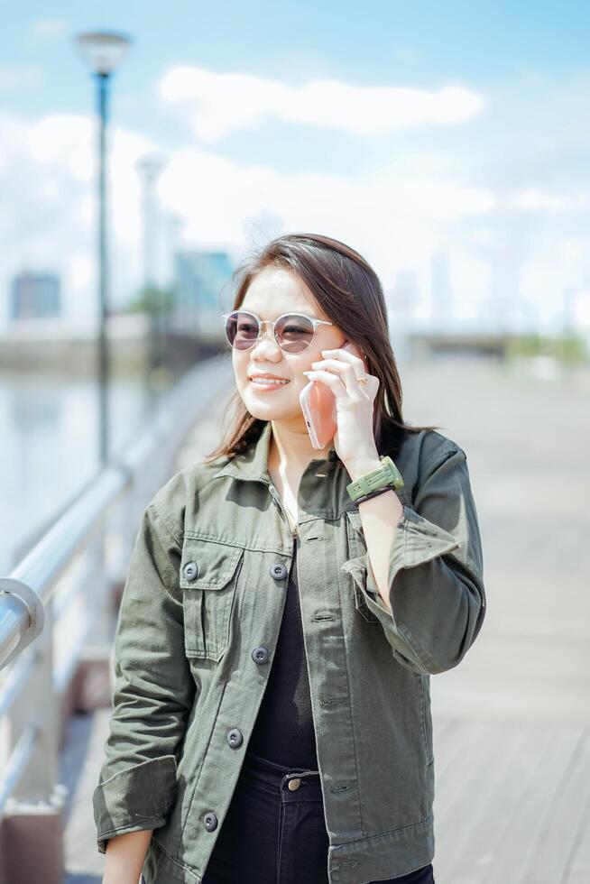 utilizando teléfono inteligente de joven hermosa asiático mujer vistiendo chaqueta y negro pantalones posando al aire libre foto