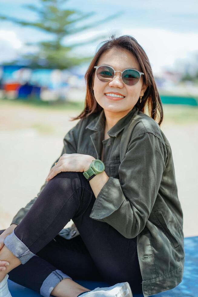 joven hermosa asiático mujer vistiendo chaqueta y negro pantalones posando al aire libre sentado en el parque foto