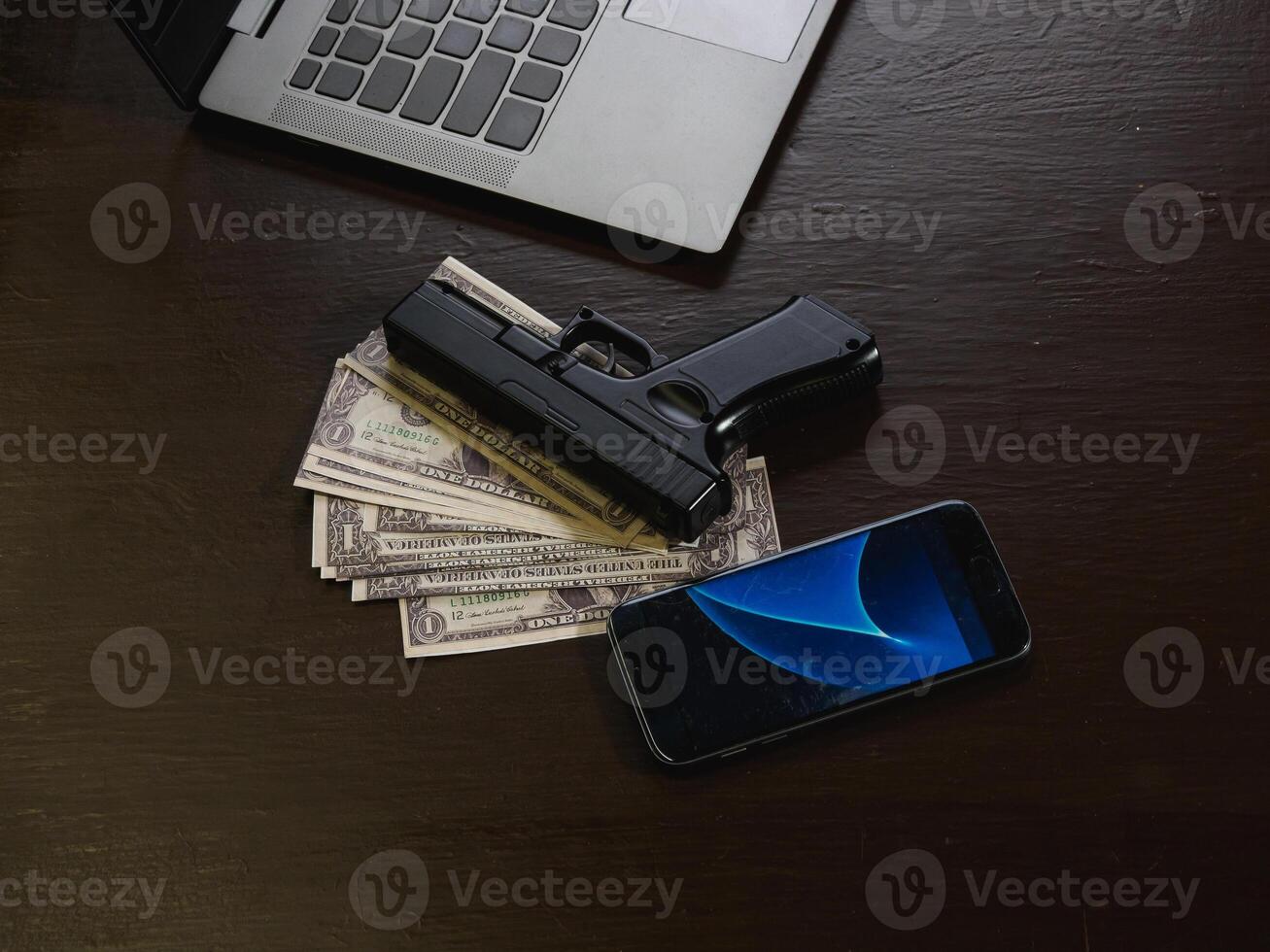 Computers, mobile phone, money, guns, and other devices that hackers use to steal or hijack information to trick victims into taking money. laying on wooden table in dark room in mysterious apartment. photo