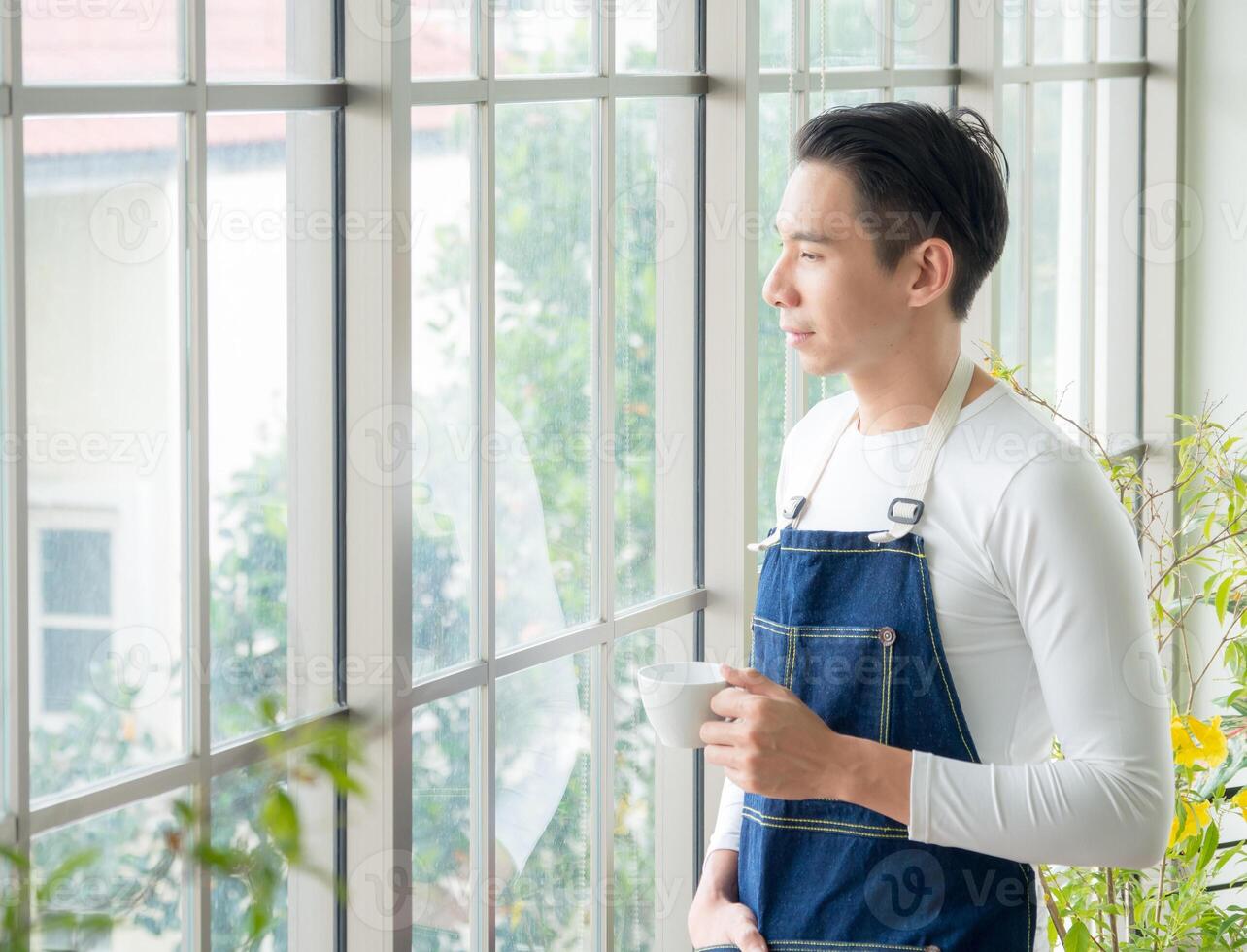 Portrait gardener adult young Asian man one person handsome standing think look and Smile relax happy hand holding drinking cup coffee in room cafe with glass window and green tree in morning time. photo
