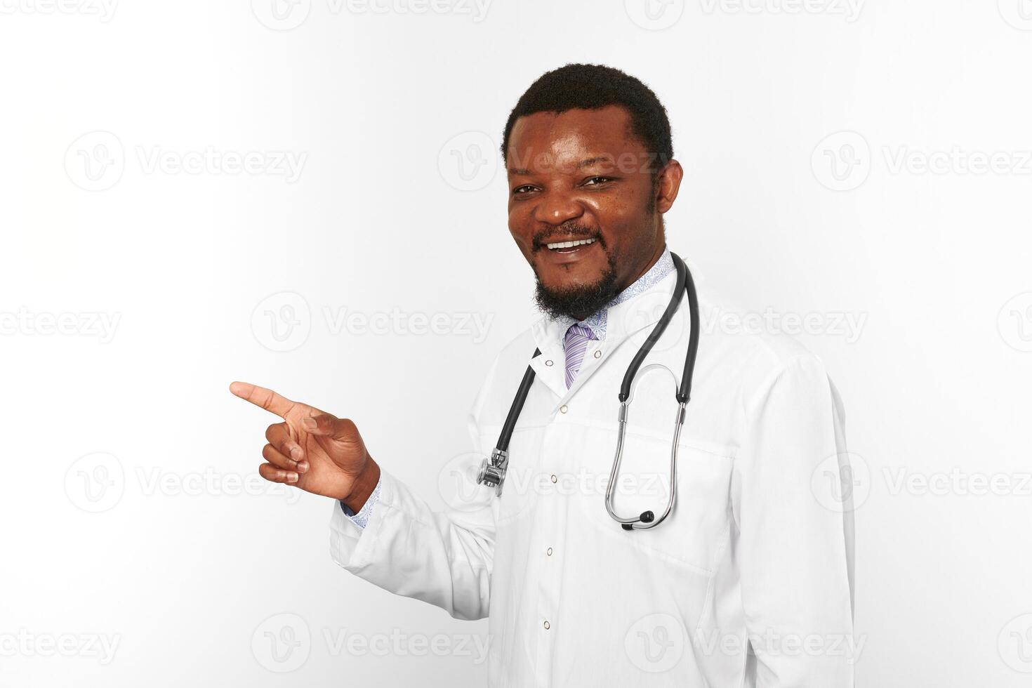 Smiling black bearded doctor man in white coat with stethoscope points finger, white background photo