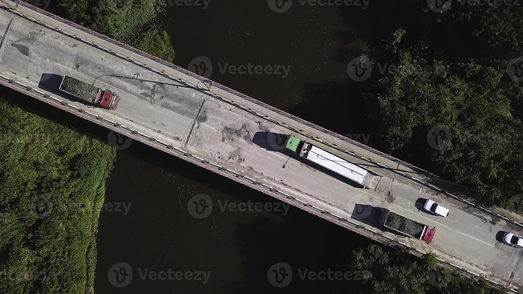 Top view of passing vehicles on bridge over river. Clip. Trucks and cars drive over bridge over brown river. Driving across bridge on sunny summer day photo