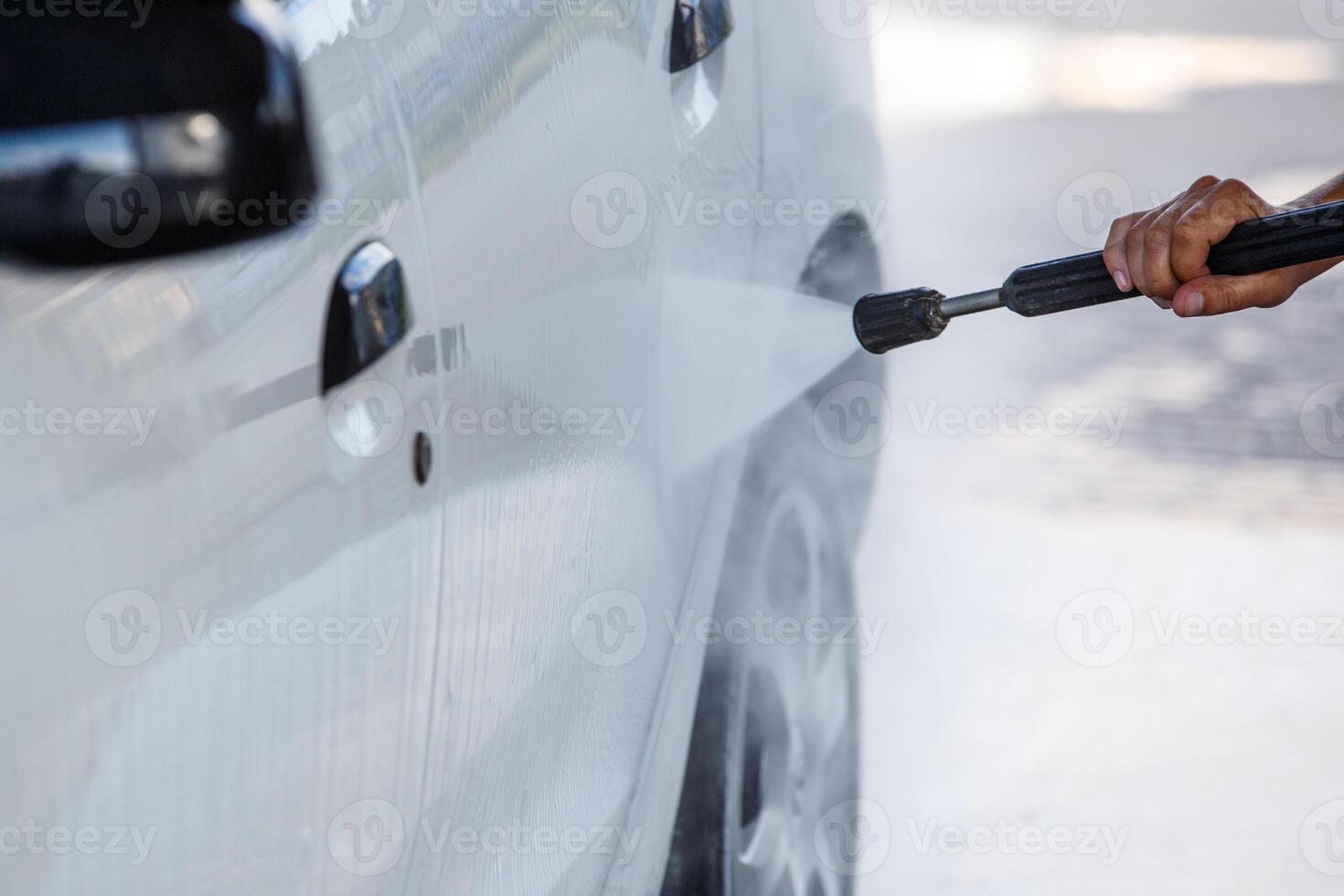 nozzle with high pressure water washer spraying white car at public self service car washing station, closeup with selective focus photo