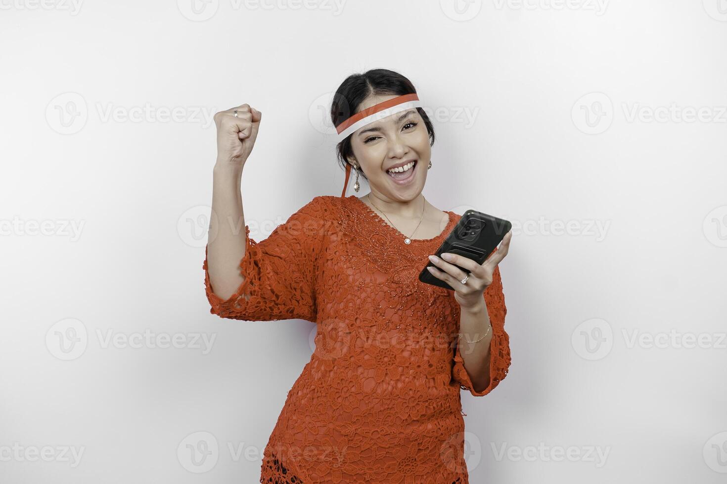 A young Asian woman with a happy successful expression while holding her phone and wearing red kebaya, flag headband isolated by white background. Indonesia's independence day concept. photo