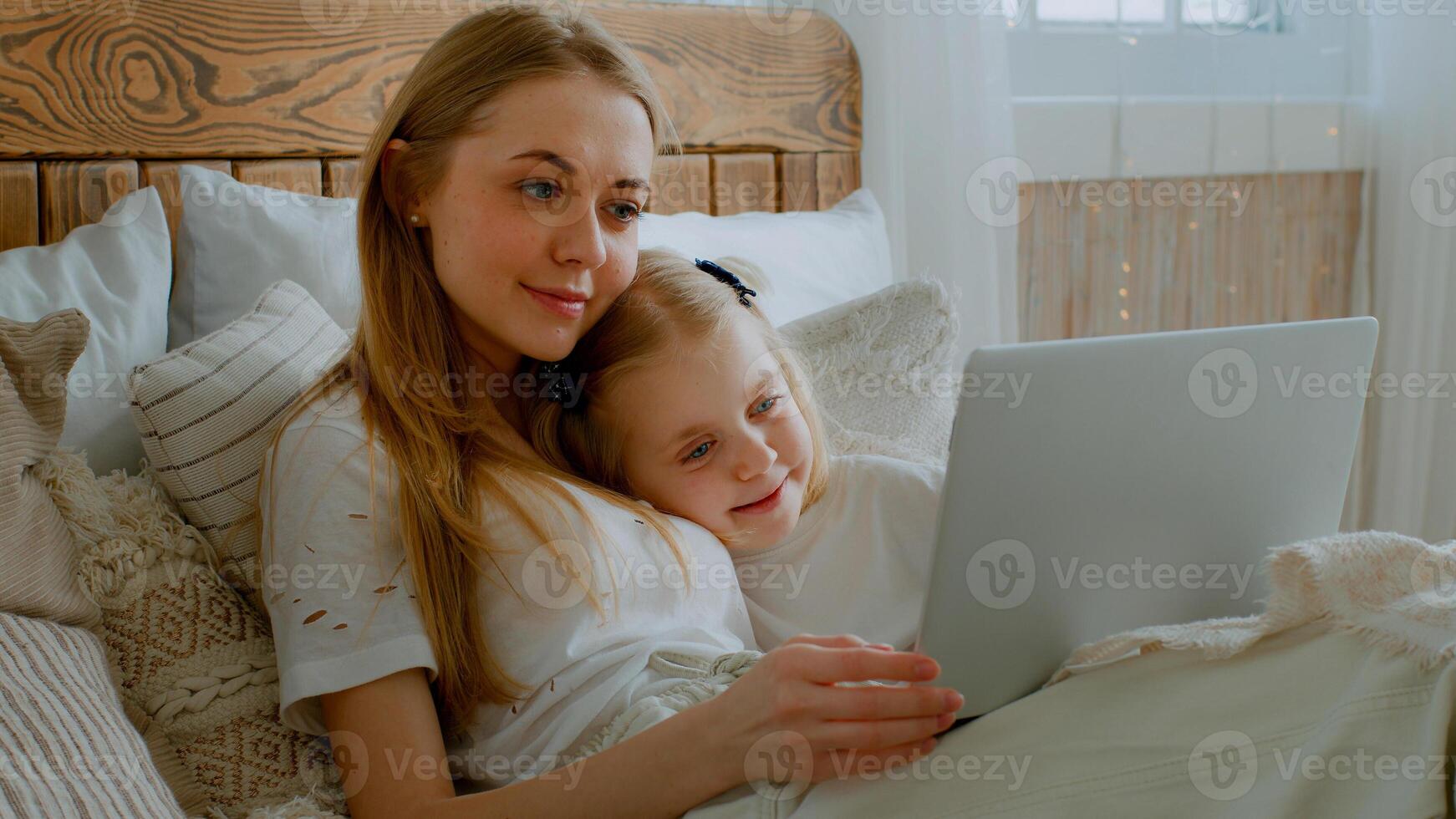 Small little daughter with Caucasian mom woman mother relax in bed together affectionate lying in bedroom with laptop video call conference talk watching movie film online cartoon in internet talking photo