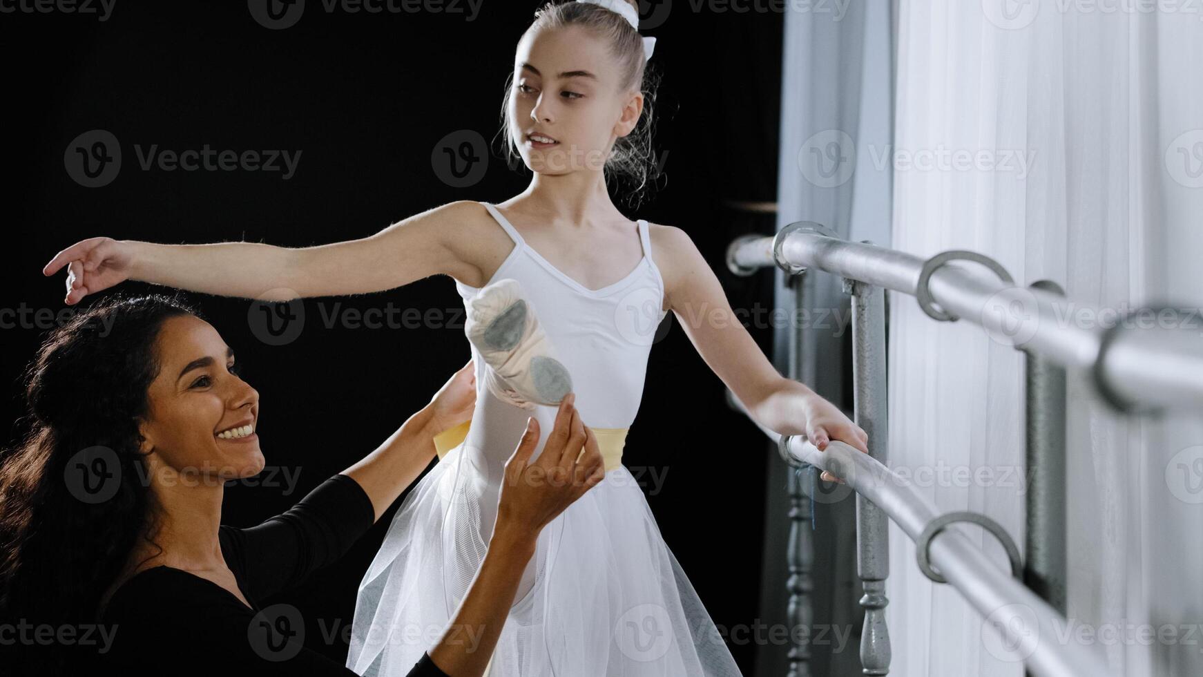Girl teenager ballerina student doing classic exercises near ballet barre trains listens to advice of woman teacher professional female trainer helps with stretching holds child's leg corrects posture photo