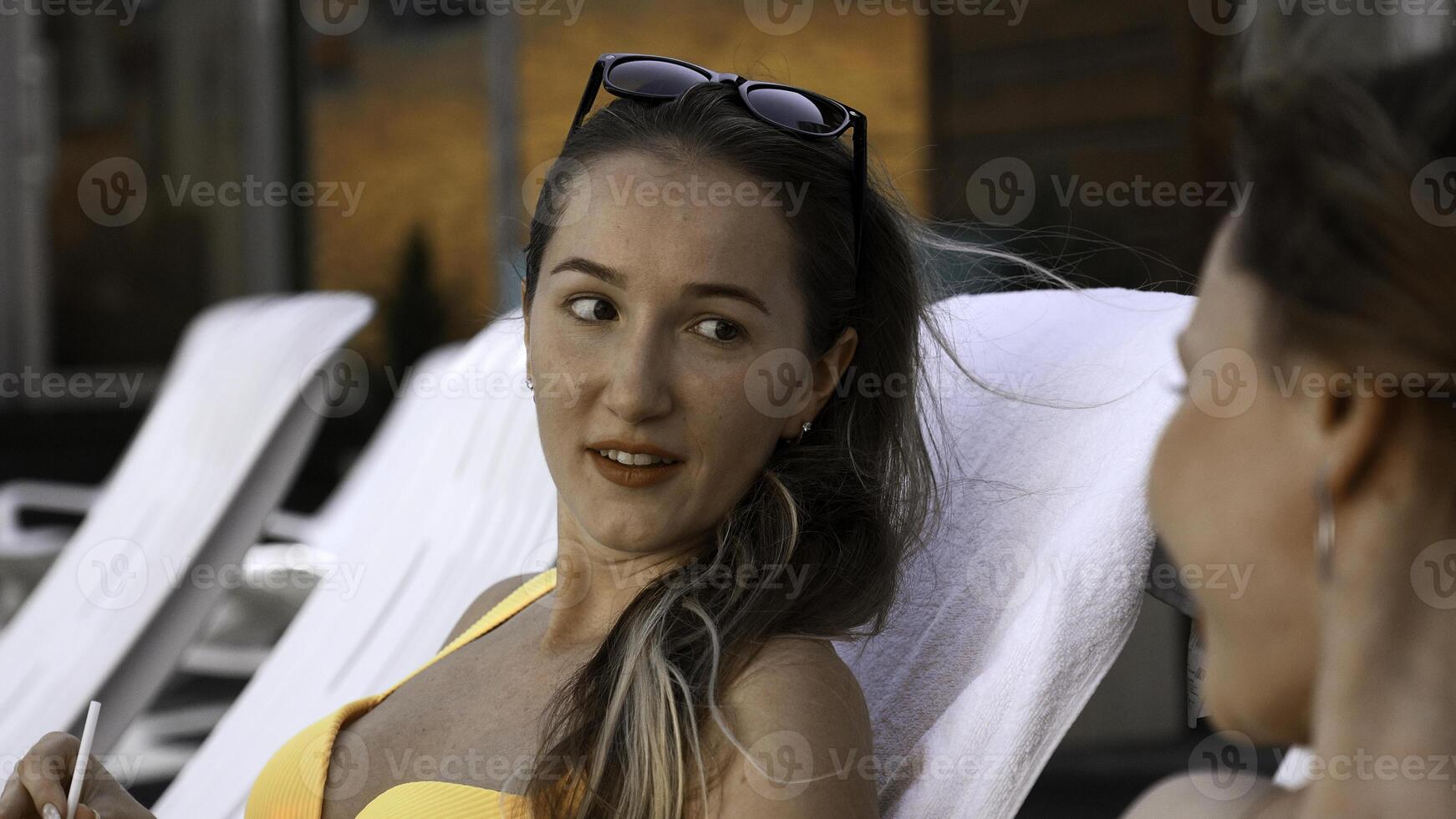 Happy young women lying on sunbeds by the swimming pool and gossiping. Clip. Relaxing near resort swimming pool with a cocktail. photo