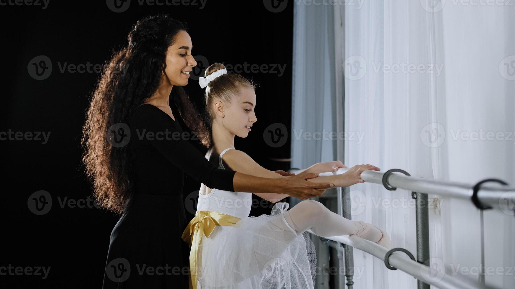 Teen girl child teenager ballerina in tutu has advice of woman coach teacher learn to dance in dancing school near ballet barre stretching leg hip. Female trainer helps with stretch correct position photo