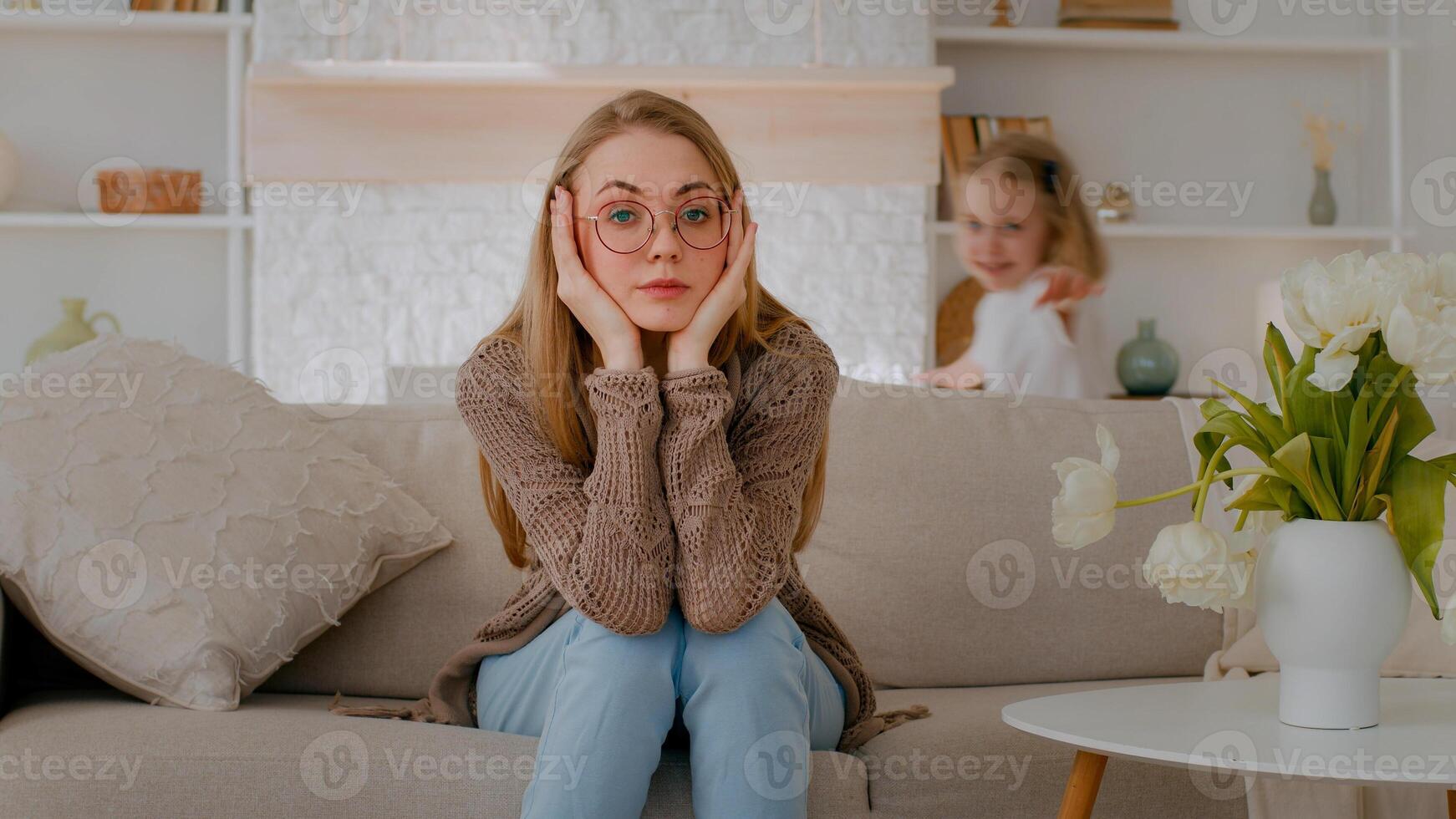 Tired upset Caucasian mother sitting on sofa at home sad frustrated stressed exhausted fatigued mom suffer headache with active noise difficult kid child baby girl daughter running around playing photo