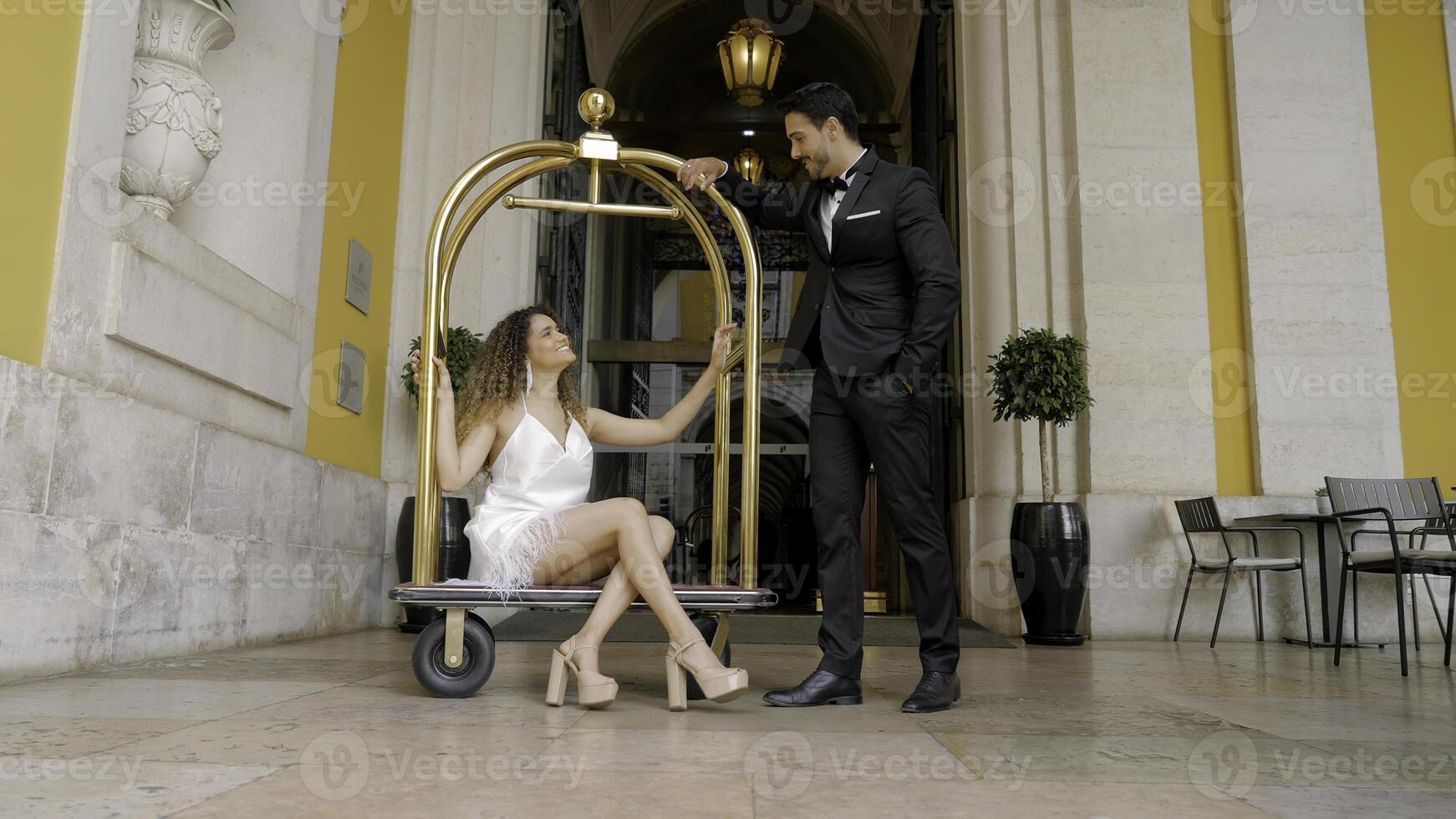 Elegant couple at entrance to hotel. Action. Elegant woman sits on European luggage trolley in hotels. Beautiful luxury couple at expensive hotel photo