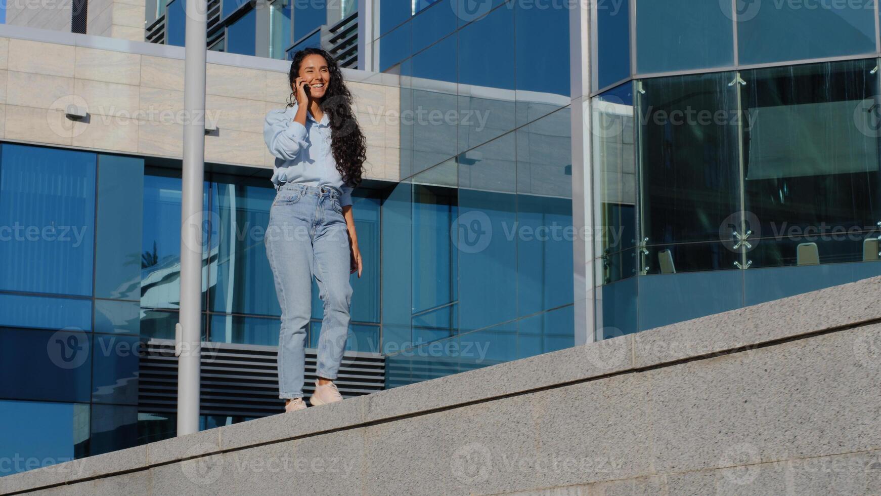 joven alegre hermosa morena negocio mujer ganador despreocupado niña caminando en urbano azul vaso edificio antecedentes hablando móvil teléfono bailando Moviente felicidad respondiendo llamada en ciudad yendo bailes foto