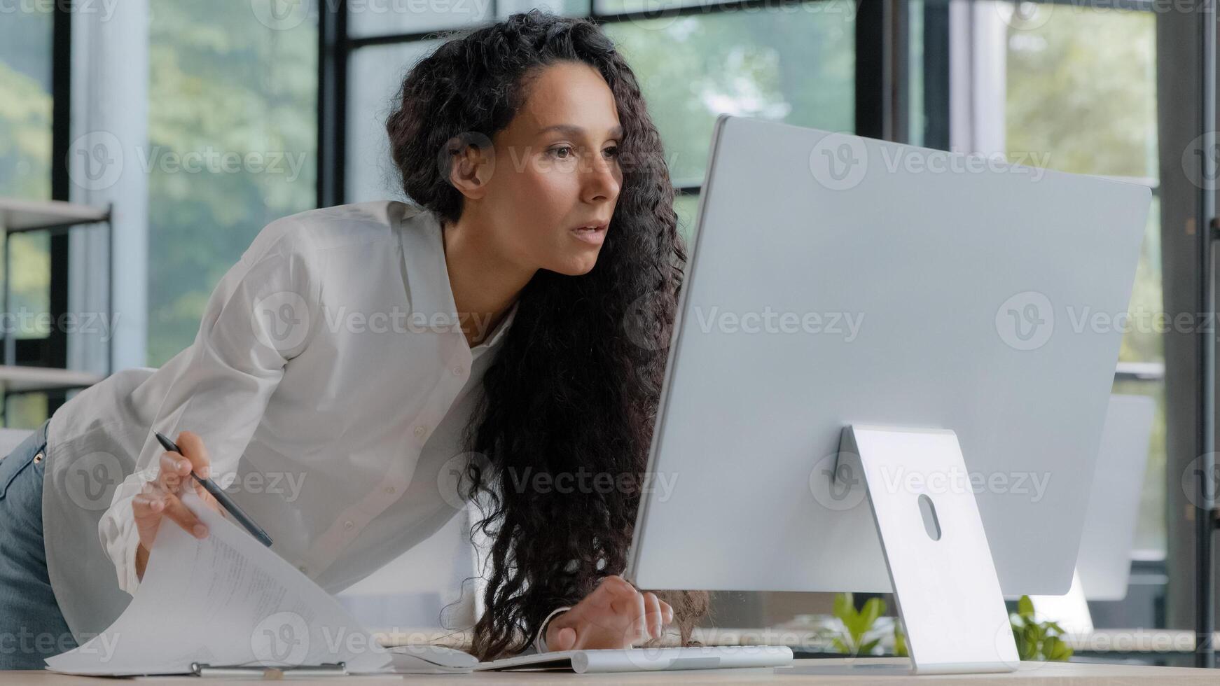 Young attractive hispanic woman professional manager developer working in modern office focused businesswoman checking electronic information on computer using paper documents researching sales data photo