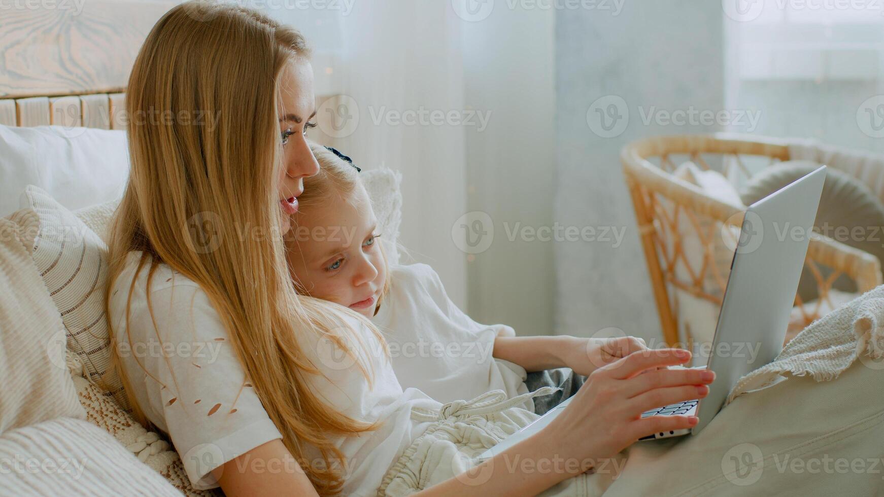 Caucasian mother with daughter browsing laptop online store lying in bed buying in internet shopping watch movie at bedroom together. Mom teach little small baby child girl using computer cyberspace photo