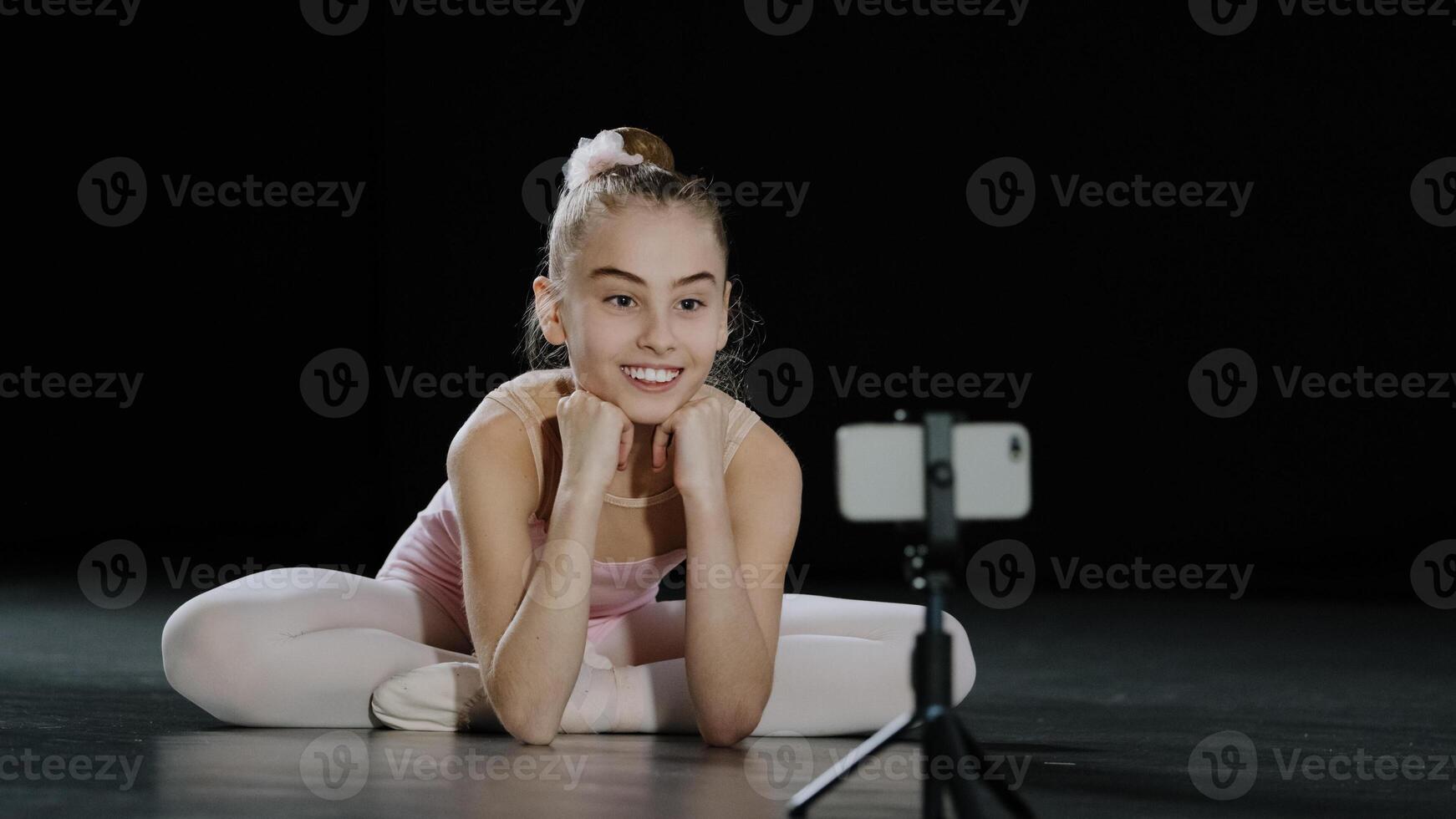 Young teen girl child teenager ballerina gymnast dancer sitting on floor recording vlog waving hello to mobile phone camera on tripod has online video call conference chat live broadcasting smartphone photo