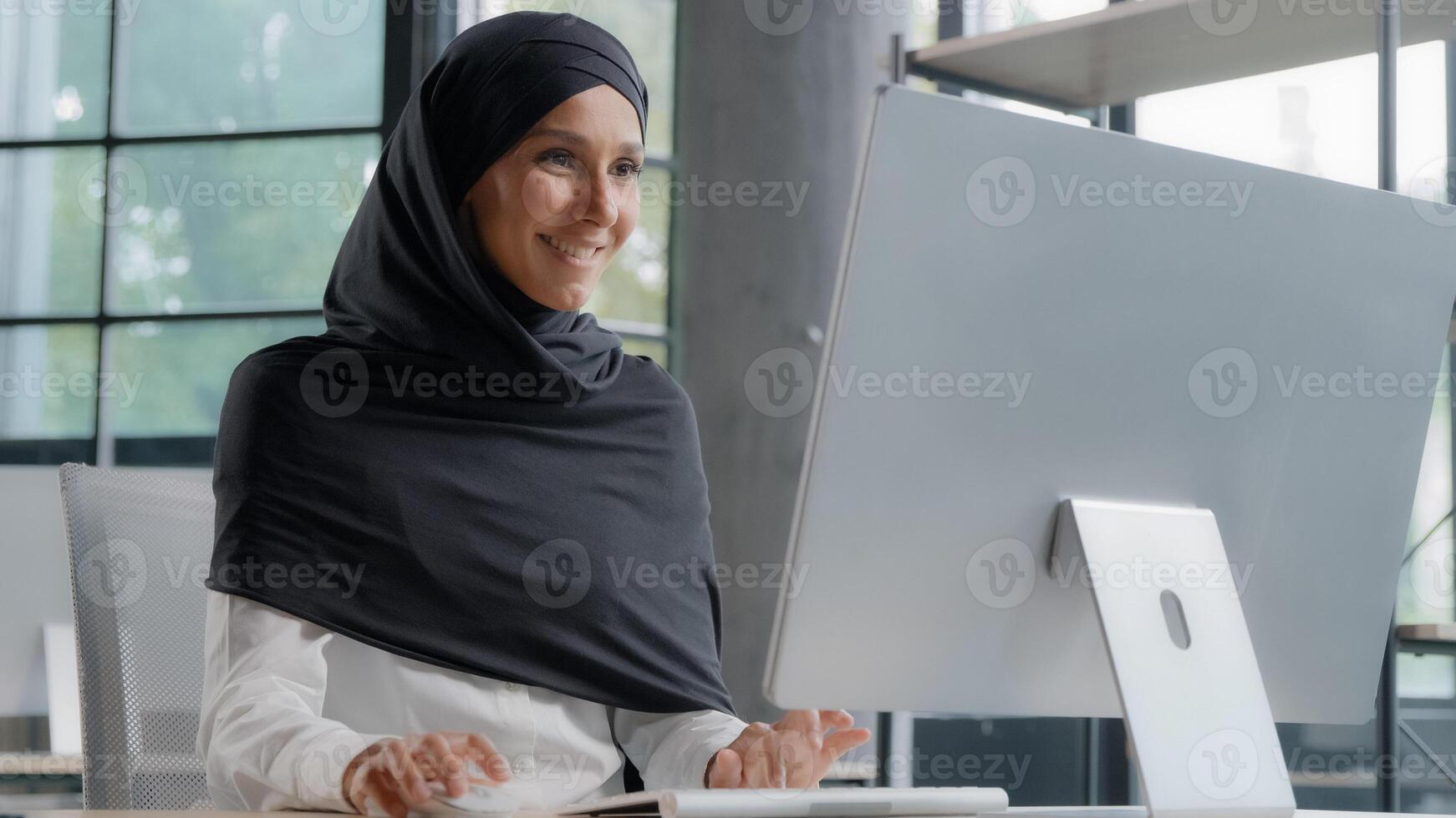 Young arab businesswoman in hijab working on computer smiling enjoying office work successful woman professional manager company employee develops project online searching for information on Internet photo