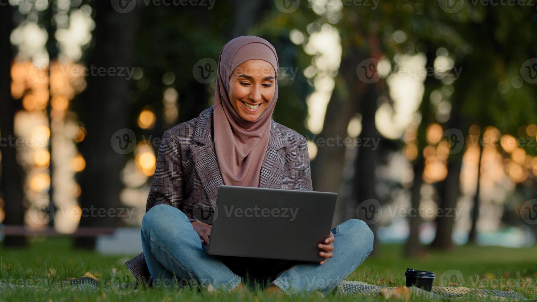 Happy smiling muslim islamic business woman girl female student wears hijab sitting on green grass lawn in city park using laptop for distant e-learning work chatting online checking mail with coffee photo