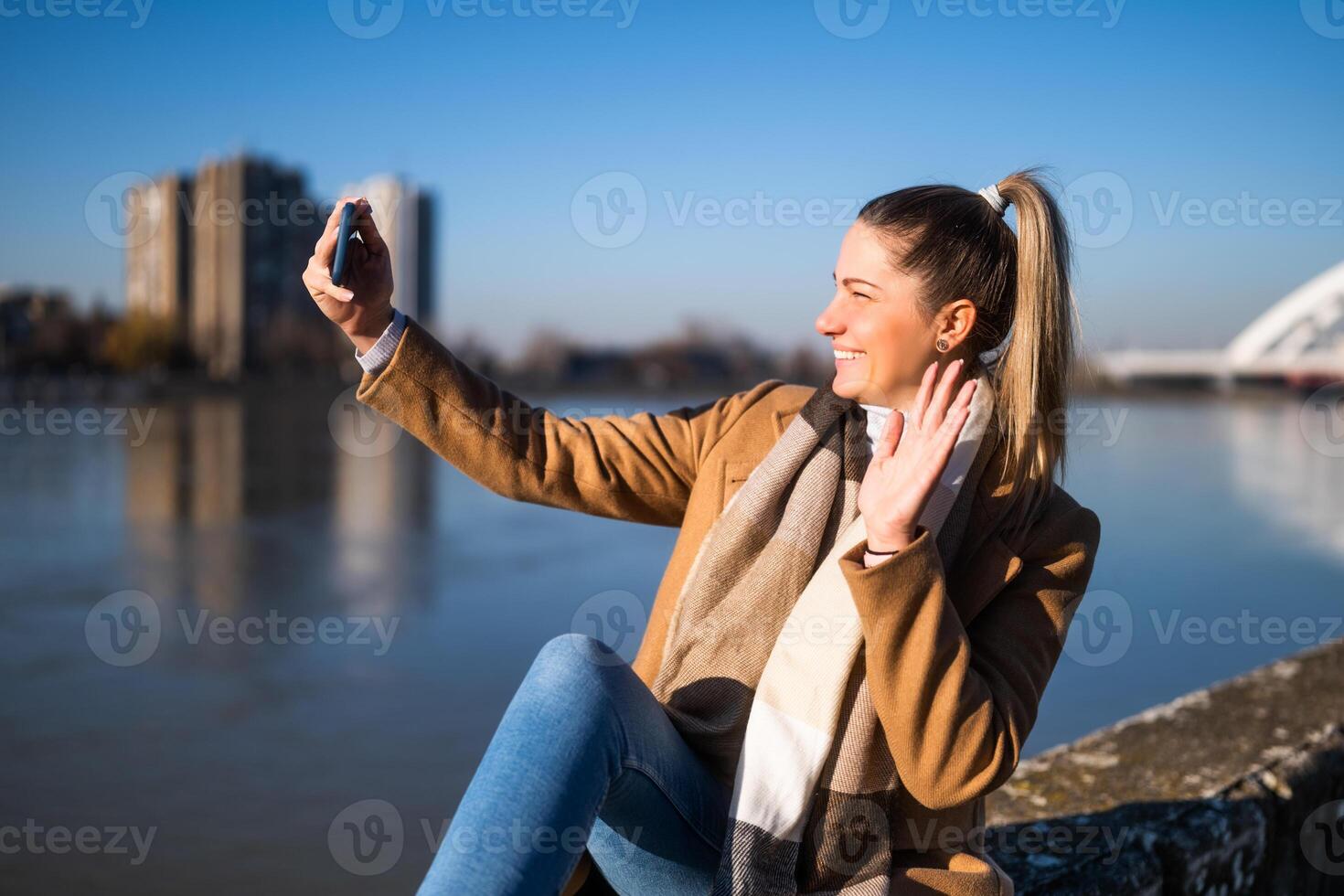 Beautiful woman in warm clothing taking selfie with phone and waving while enjoys resting by the river on a sunny winter day. photo