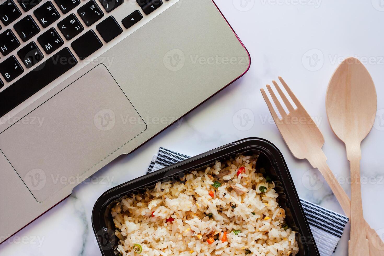 Delicious fried rice in lunch box on napery with wooden spoon and fork and computer laptop at workplace photo