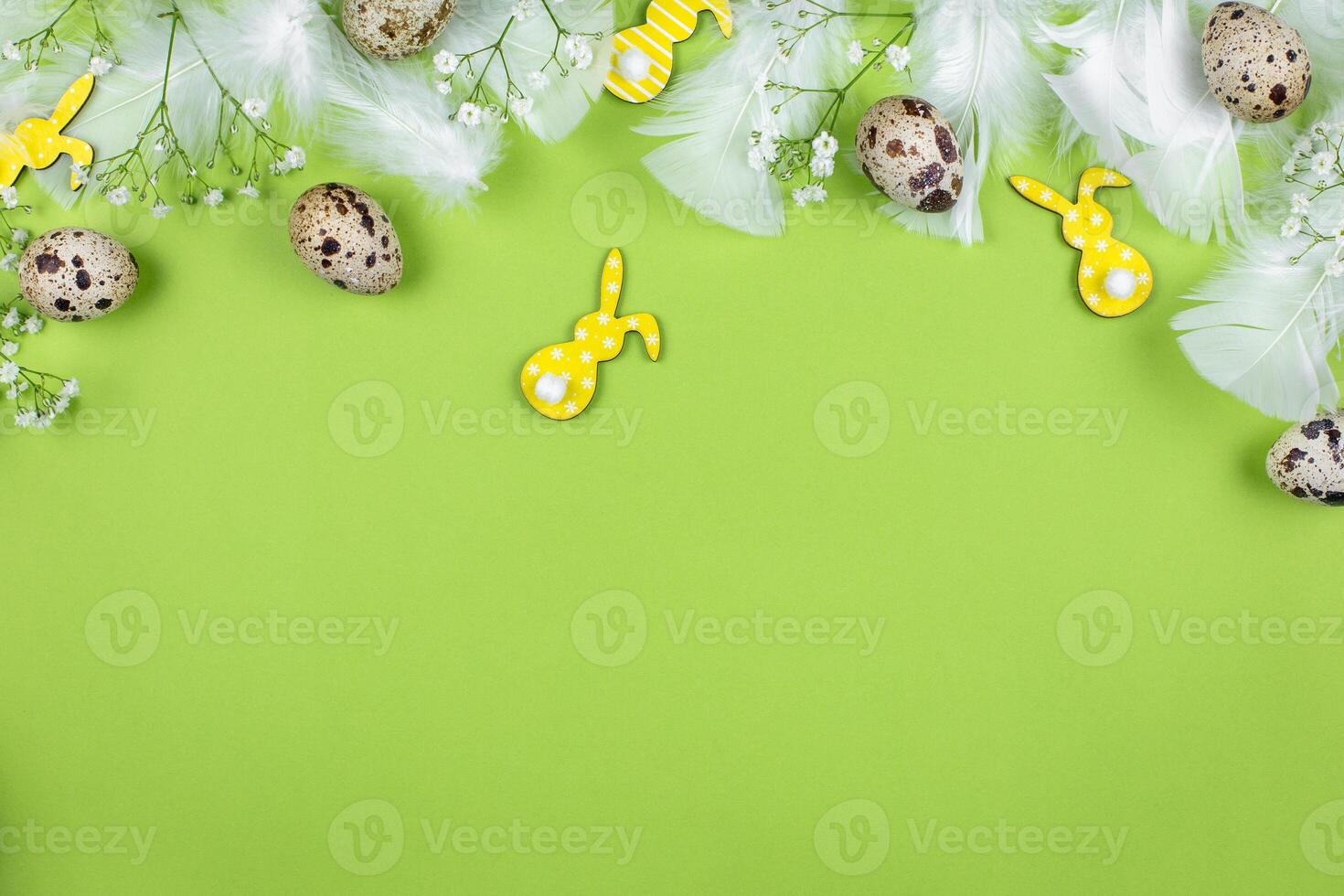 Easter border flat lay with quail eggs, spring flowers, yellow bunnies and white feathers on a green background. Top view. Copy space. photo