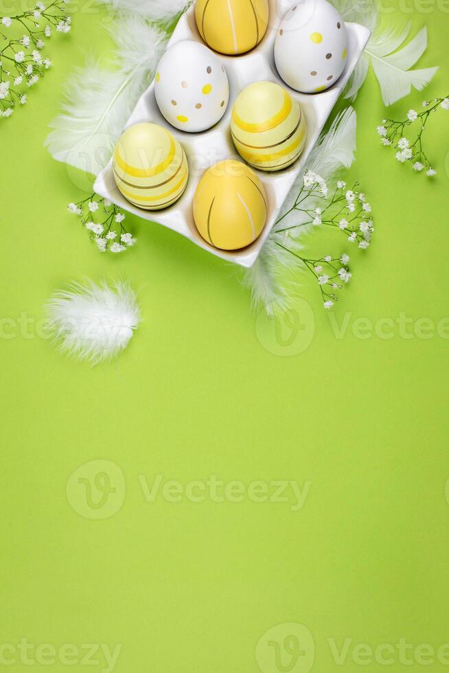 Easter flat lay with colorful eggs in a white ceramic holder, spring flowers and white feathers on a green background. Top view. Copy space. photo