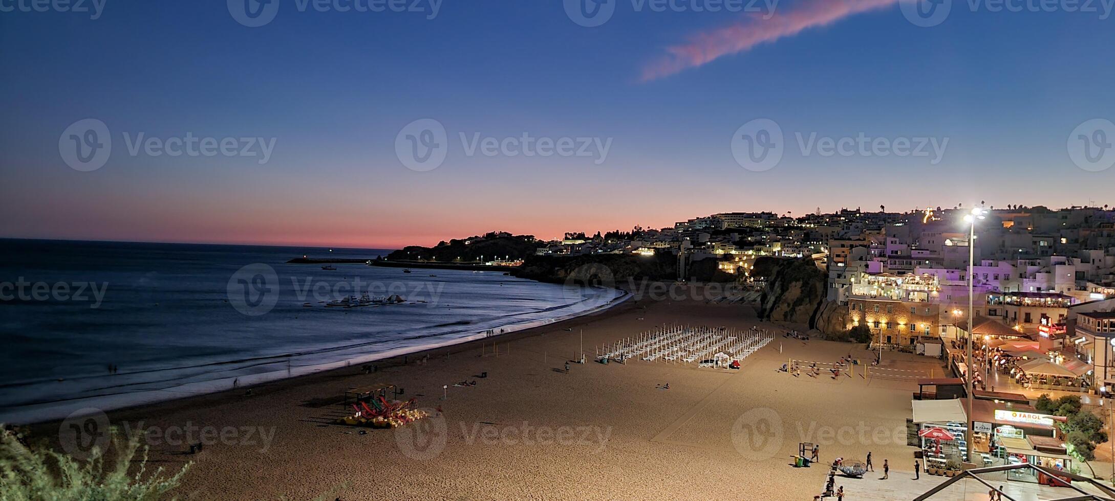 Panoramic view of a coastal town at dusk with illuminated streets, a serene beach, and a tranquil sea under a gradient sky. photo