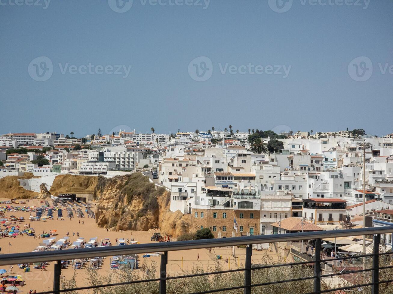 Scenic view of a coastal town with white buildings, sandy beach, and cliffs under a clear blue sky. photo
