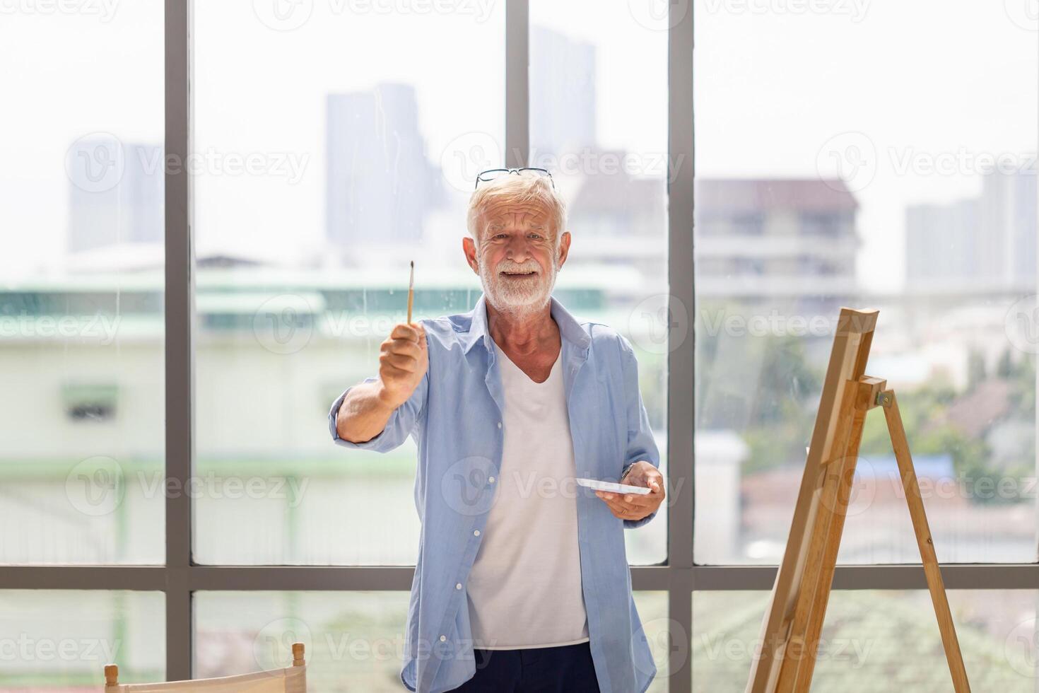 Cheerful senior man showing paintbrush and painting on canvas at home, Elderly man painting on a canvas, Happy retirement concepts photo