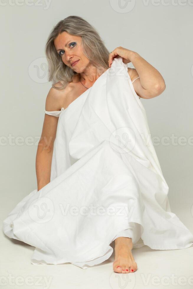 un hermosa mayor mujer con gris pelo envuelto en un blanco sábana poses en contra un gris antecedentes. foto