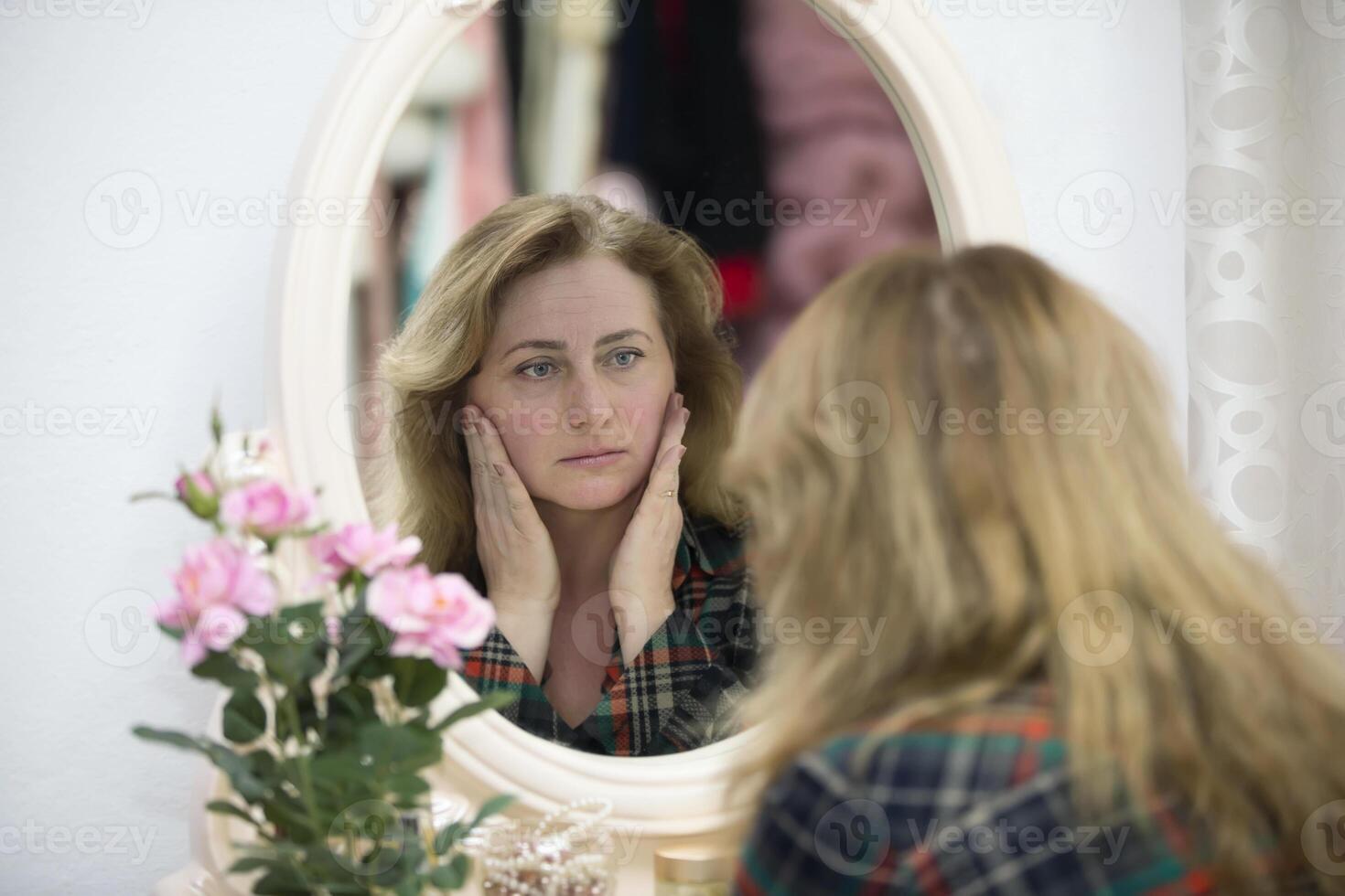 An elderly woman looks in the mirror at the wrinkles. Woman touches her aged face photo