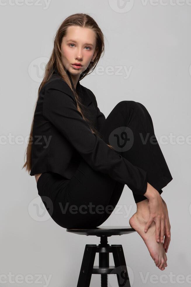 A slender beautiful teenage girl in a black jacket sat in a beautiful pose on a studio chair. Blonde fashion model girl posing. The dream is to be a model. photo